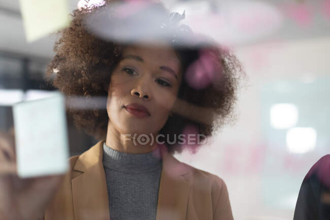 Klug gekleidete afrikanisch-amerikanische Geschäftsfrau schreibt im Büro Notizen an eine Glaswand. unabhängiges kreatives Designgeschäft. — Stockfoto