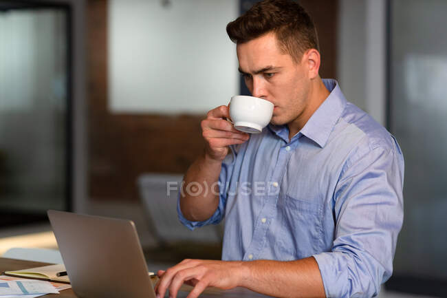 Uomo d'affari caucasico occasionale che usa il computer, beve caffè e si siede alla scrivania. uomo d'affari al lavoro in ufficio moderno. — Foto stock
