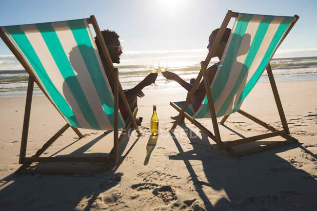 Casal afro-americano apaixonado sentado em espreguiçadeiras, apreciando bebidas na praia. amor, romance e férias de verão na praia. — Fotografia de Stock