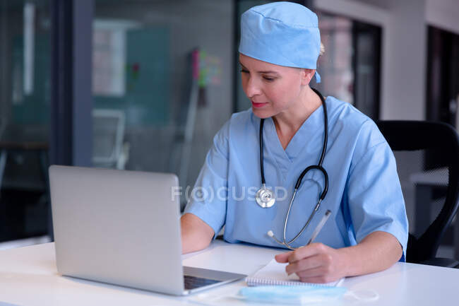 Kaukasische Ärztin am Schreibtisch mit einem Laptop, der Notizen während eines Videogesprächs macht. Telemedizin während der Quarantäne. — Stockfoto