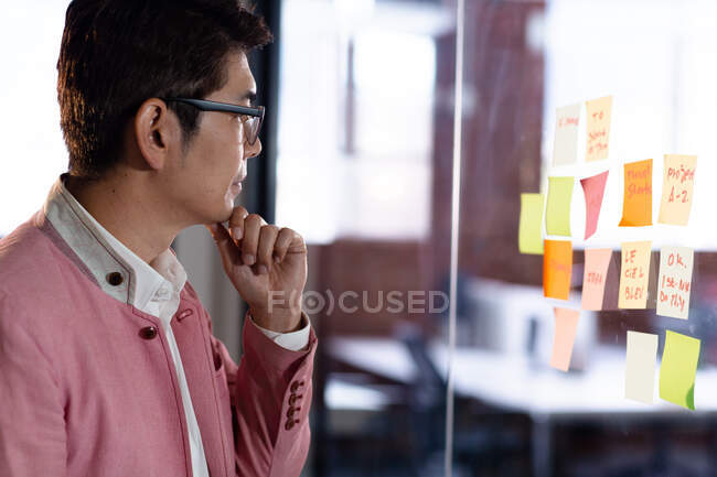 Stylish asian businessman reading memo notes on glass wall. business person at work in modern office. — Stock Photo