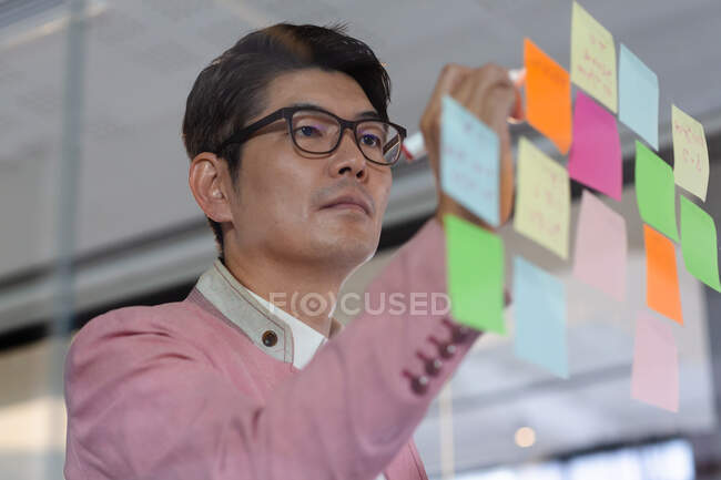 Stylish asian businessman writing notes on glass wall. business person at work in modern office. — Stock Photo