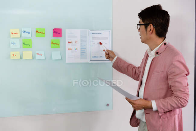Stilvoller asiatischer Geschäftsmann, der Zettel mit Dokumenten an einer Glaswand hält. Geschäftsmann bei der Arbeit im modernen Büro. — Stockfoto