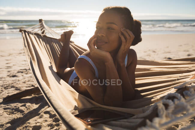 Happy african american woman lying in hammock on beach looking ahead. healthy outdoor leisure time by the sea. — Stock Photo