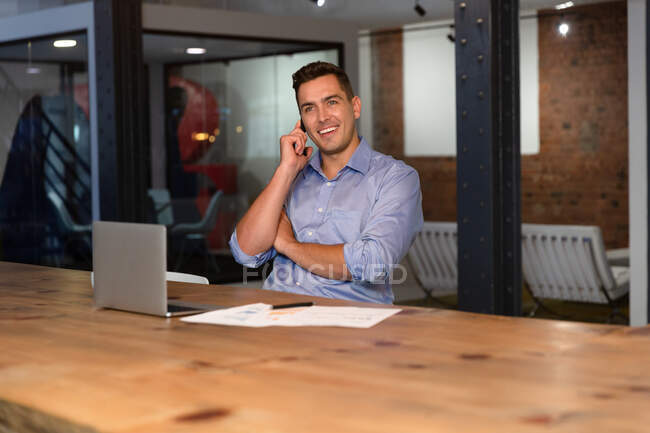 Portrait of happy casual caucasian businessman talking on smartphone sitting at desk. business person at work in modern office. — Stock Photo