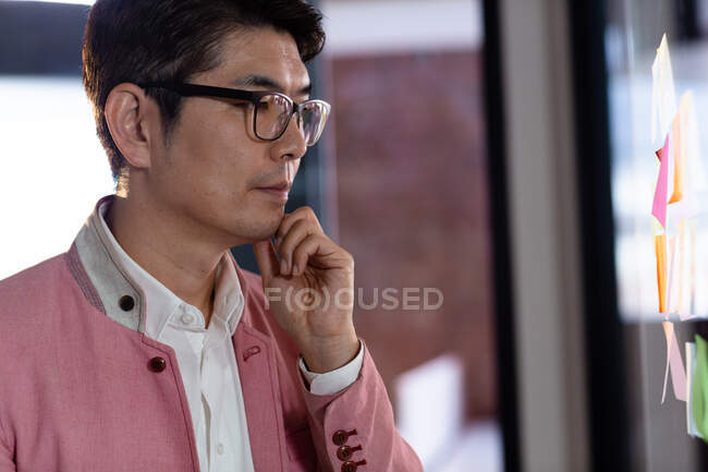 Стильный азиатский бизнесмен, читающий заметки на стеклянной стене. деловой человек за работой в современном офисе. — стоковое фото