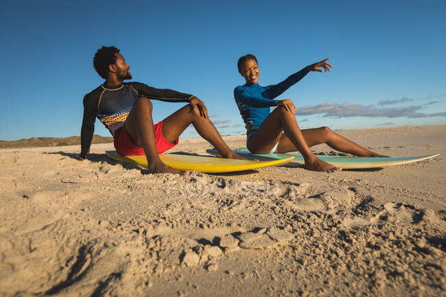 Счастливая африканская американская пара на пляже, сидящая на досках для сёрфинга, указывая. здоровый отдых на открытом воздухе у моря. — стоковое фото