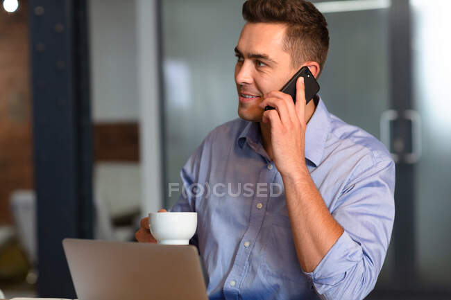 Heureux homme d'affaires caucasien occasionnel parlant sur smartphone boire du café assis au bureau. homme d'affaires au travail dans un bureau moderne. — Photo de stock