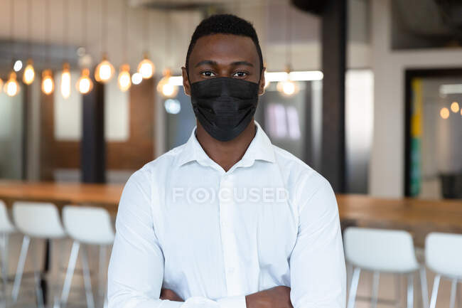 Retrato de un hombre de negocios afroamericano casual con máscara facial y mirando a la cámara. persona de negocios en el trabajo en la oficina moderna durante covid 19 pandemia coronavirus. - foto de stock