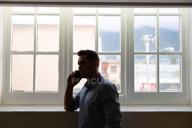 Homem de negócios caucasiano elegante em pé à janela e falando no smartphone. pessoa de negócios no trabalho em escritório moderno. — Fotografia de Stock