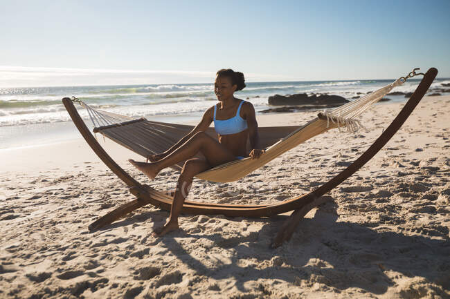 Mujer afroamericana feliz sentada en hamaca en la playa. tiempo de ocio al aire libre saludable junto al mar. - foto de stock