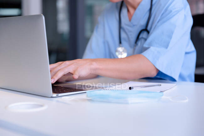 Midsection of caucasian female doctor sitting at desk using laptop computer. profesional médico en el trabajo. - foto de stock