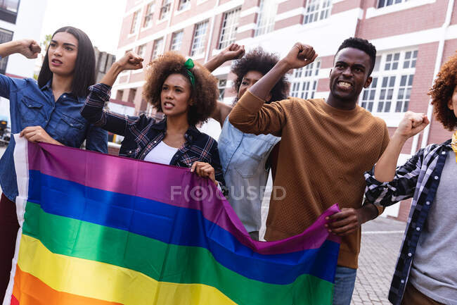 Diversos manifestantes masculinos y femeninos en marcha sosteniendo la bandera del arco iris, gritando y levantando puños. marcha por la igualdad de derechos y justicia. - foto de stock