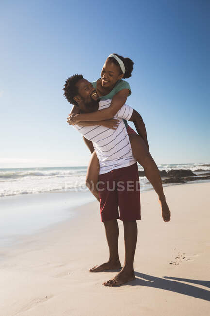 Una feliz pareja afroamericana en la playa se miran a cuestas. tiempo de ocio al aire libre saludable junto al mar. - foto de stock