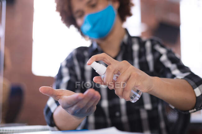 Empresario mixto en la oficina con máscara desinfectando manos. negocio creativo independiente durante la pandemia de coronavirus covid 19. - foto de stock