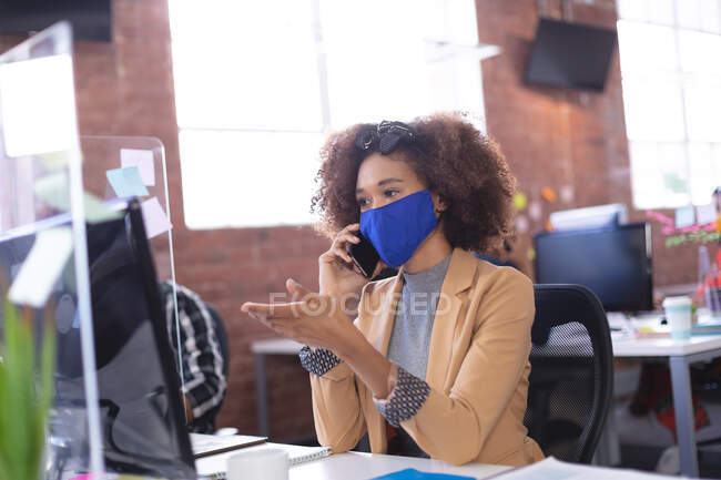 Afroamerikanische Geschäftsfrau mit Maske spricht im Büro per Smartphone. unabhängiges kreatives Design-Geschäft während der Covid 19 Coronavirus-Pandemie. — Stockfoto