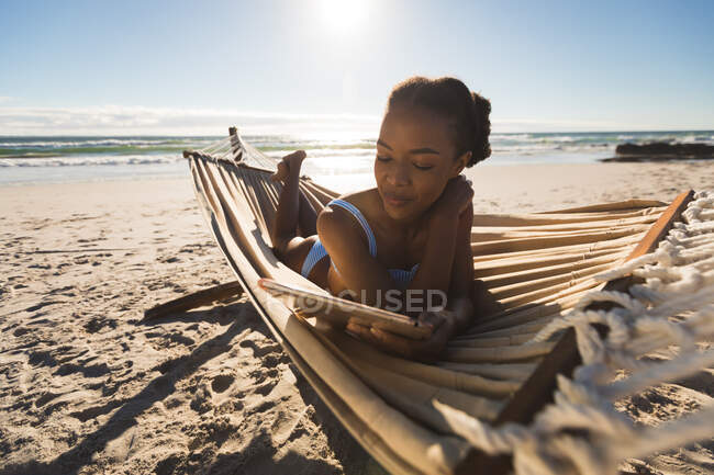 Feliz mulher afro-americana deitada em rede na praia usando tablet. tempo de lazer ao ar livre saudável pelo mar. — Fotografia de Stock