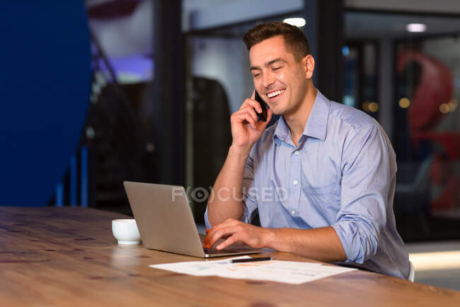 Portrait of happy casual caucasian businessman talking on smartphone at desk using laptop. business person at work in modern office. — Stock Photo
