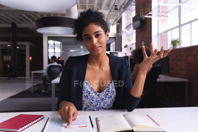 Femme d'affaires métisse assise au bureau avec des documents montrant un geste d'appel vidéo. entreprise indépendante de design créatif. — Photo de stock