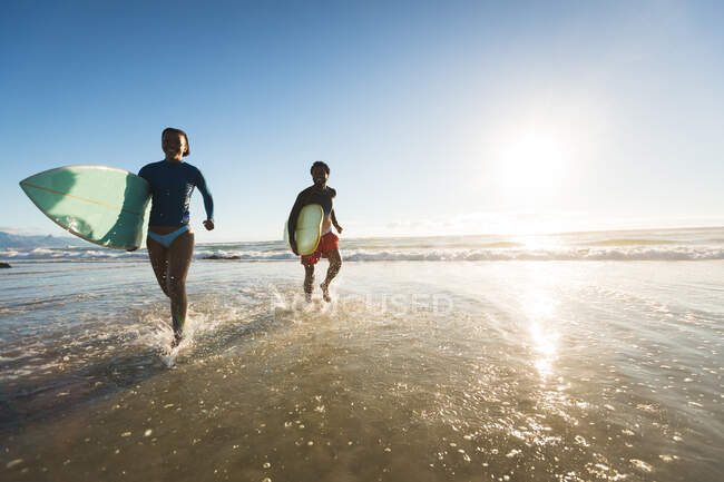 Happy african american couple running out the sea carrying surfboards. healthy outdoor leisure time by the sea. — Stock Photo