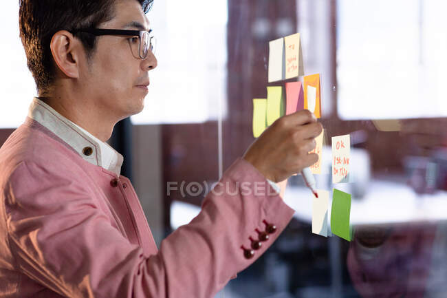 Lächelnder asiatischer Geschäftsmann klebt Notizen an Glaswand. Geschäftsmann bei der Arbeit im modernen Büro. — Stockfoto