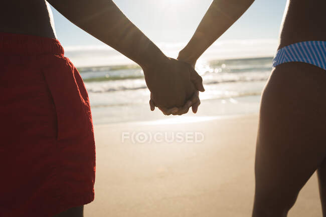 Мидсекция африканской американской пары, влюбленной в друг друга, держится за руки. здоровый отдых на открытом воздухе у моря. — стоковое фото