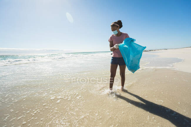 Afroamerikanerin mit Gesichtsmaske sammelt Müll am Strand. Öko-Strandschutz während der Coronavirus-Pandemie. — Stockfoto