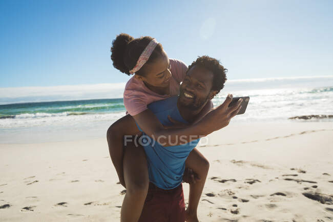 Счастливая африканская американская пара на пляже у моря, катается на спине и делает селфи. здоровый отдых на открытом воздухе у моря. — стоковое фото