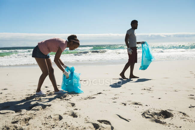 African american couple wearing latex gloves collecting rubbish from the beach. eco summer beach conservation — Stock Photo