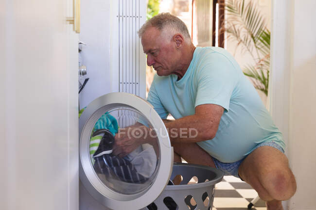 Kaukasischer älterer Mann hockt an der Waschmaschine und holt Wäsche. Isolationshaft während der Quarantäne. — Stockfoto