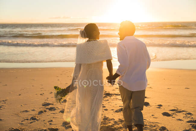 Pareja afroamericana enamorada de casarse, caminando por la playa al atardecer tomados de la mano. amor, romance y boda vacaciones de verano vacaciones de playa. - foto de stock