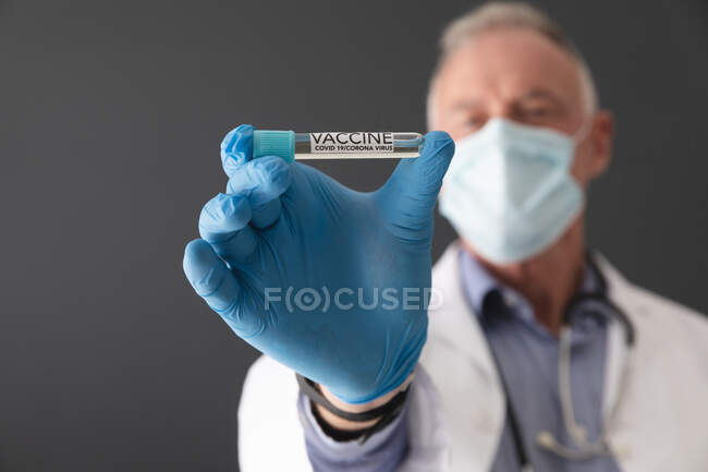 Caucasian senior male doctor wearing face mask and surgical gloves holding vial of covid 19 vaccine. medical professional at work during coronavirus covid 19 pandemic. — Stock Photo