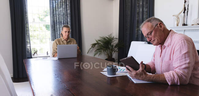 Кавказская старшая пара в гостиной сидит за столом и работает, используя ноутбук и планшет. оставаться дома в изоляции во время карантинной изоляции. — стоковое фото