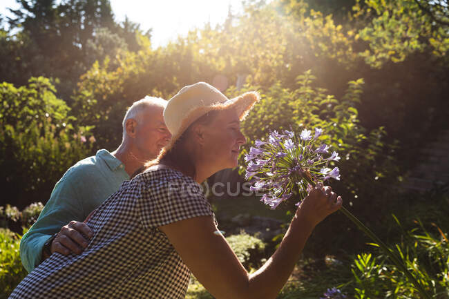 Glückliche kaukasische Senioren spazieren im sonnigen Garten und riechen Blumen. Isolationshaft während der Quarantäne. — Stockfoto