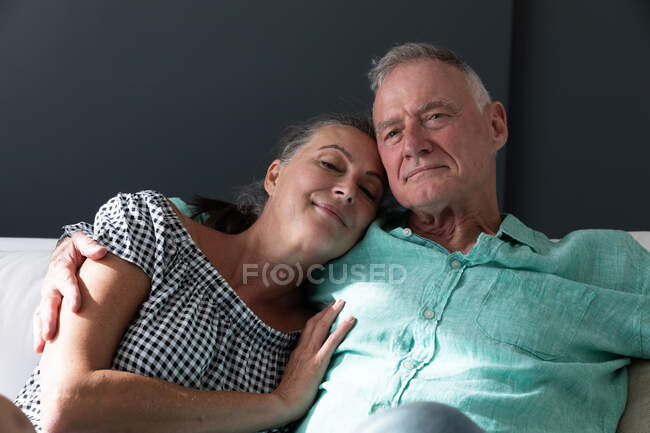 Счастливая старшая кавказская пара отдыхает в гостиной, сидя на диване обнимаясь и улыбаясь. оставаться дома в изоляции во время карантинной изоляции. — стоковое фото
