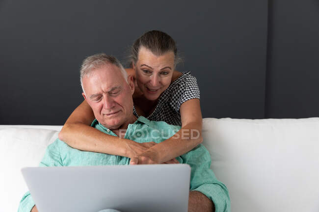 Счастливая старшая белая пара в гостиной смотрит на ноутбук, обнимающий и улыбающийся. оставаться дома в изоляции во время карантинной изоляции. — стоковое фото
