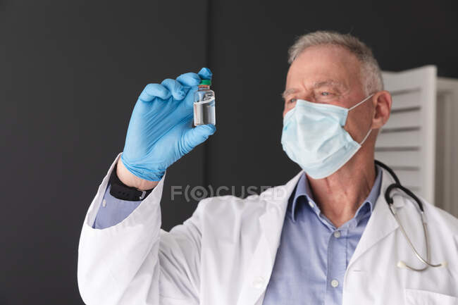 Médico varón mayor caucásico que usa mascarilla facial y guantes quirúrgicos con vial de vacuna covid 19. profesional médico en el trabajo durante la pandemia del coronavirus covid 19. - foto de stock