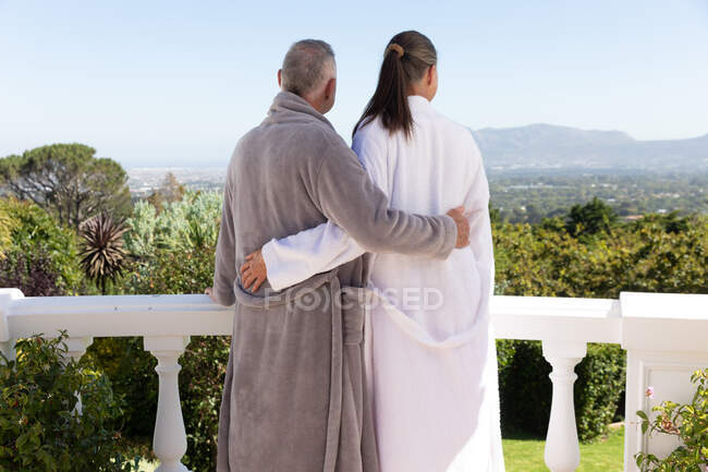 Rückansicht eines kaukasischen Seniorenpaares in Bademänteln, das sich auf dem Balkon umarmt und die Aussicht genießt. Isolationshaft während der Quarantäne. — Stockfoto