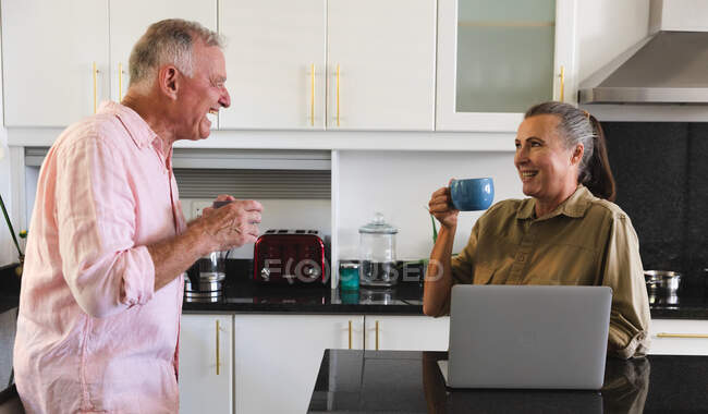 Heureux couple de personnes âgées caucasiennes dans la cuisine boire du café et rire, femme en utilisant un ordinateur portable. rester à la maison dans l'isolement pendant le confinement en quarantaine. — Photo de stock