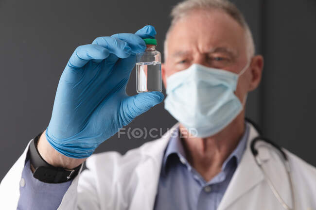 Старший врач-европеец в маске и хирургических перчатках с пузырьком с вакциной против ковида 19. медицинский работник на работе во время пандемии коронавируса. — стоковое фото