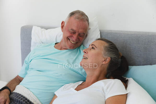 Heureux couple de personnes âgées caucasiennes se relaxant dans la chambre, s'asseyant au lit et se souriant mutuellement. rester à la maison dans l'isolement pendant le confinement en quarantaine. — Photo de stock