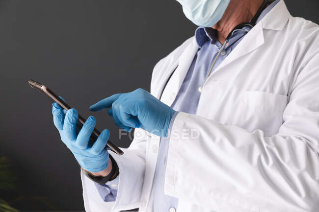 Пошкодження кавказького старшого лікаря, який носить маску для обличчя та хірургічні рукавички за допомогою планшета. медичний працівник на роботі під час пандемії коронавірусу . — стокове фото