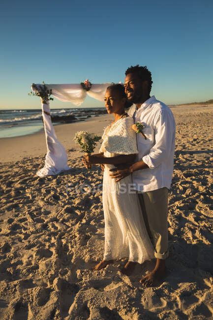 Pareja afroamericana enamorada de casarse, abrazándose en la playa mirando hacia el mar. amor, romance y boda vacaciones de verano vacaciones de playa. - foto de stock