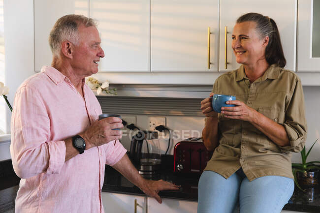 Счастливая старшая кавказская пара на кухне пьет кофе и разговаривает. оставаться дома в изоляции во время карантинной изоляции. — стоковое фото