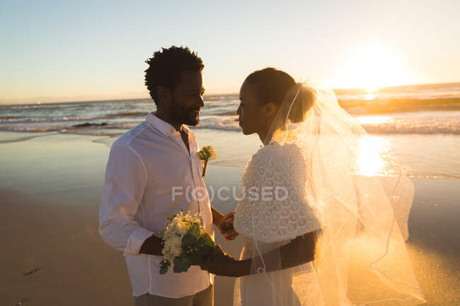 Счастливая африканская американская пара влюблённая выходит замуж, держась за руки на пляже во время заката. любовь, романтика и свадебный пляжный отдых. — стоковое фото