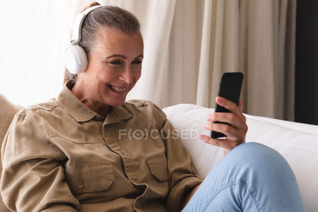 Lächelnde kaukasische Seniorin, die mit Kopfhörern auf dem Sofa sitzt und ihr Smartphone benutzt. Isolationshaft während der Quarantäne. — Stockfoto