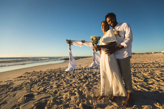 African american couple in love getting married, embracing on beach looking to camera. love, romance and wedding beach break summer holiday. — Stock Photo