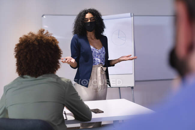 Mujer de negocios afroamericana con máscara dando presentación a diversos grupos de colegas. negocio de diseño creativo independiente. durante la pandemia de coronavirus del covidio 19. - foto de stock