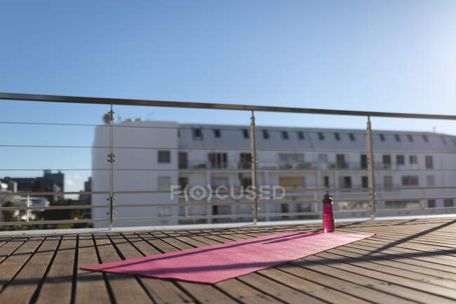Yoga mat and water bottle on sunny wooden deck of city roof terrace. staying at home in isolation during quarantine lockdown. — Stock Photo