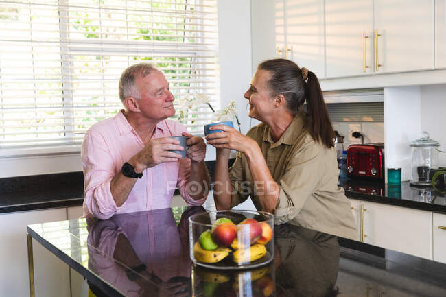 Счастливая старшая белая пара на кухне, сидящая на острове, пьет кофе и разговаривает. оставаться дома в изоляции во время карантинной изоляции. — стоковое фото